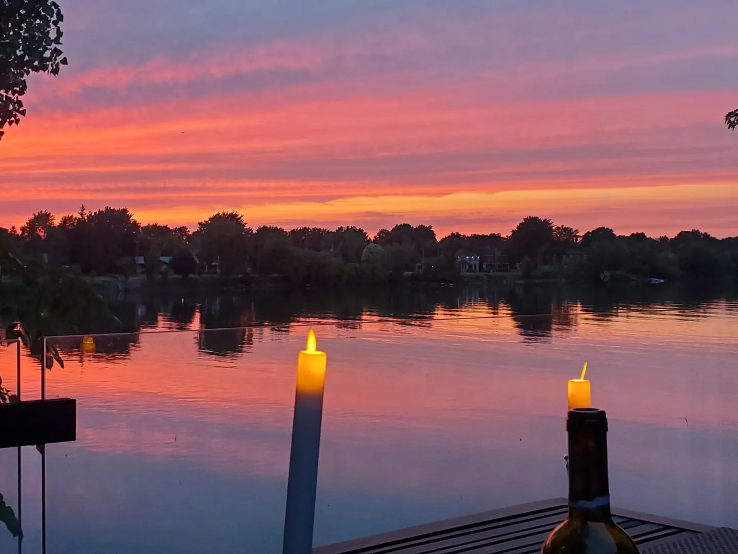 Sunset view over water with trees, houses, and two candles, one on a wine bottle, on a deck. The sky is pink, orange, and purple.