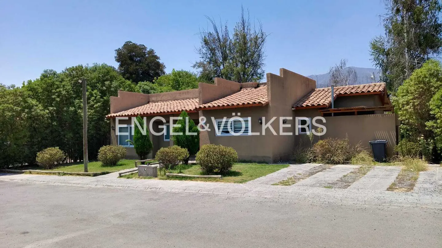 Single-story tan house with a terracotta tile roof, surrounded by green trees and shrubs under a clear blue sky.