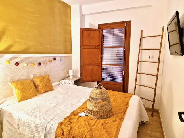Bedroom with white bedding, yellow accents, and a wooden ladder. A woven basket sits on the bed. A window with blinds is partially open.