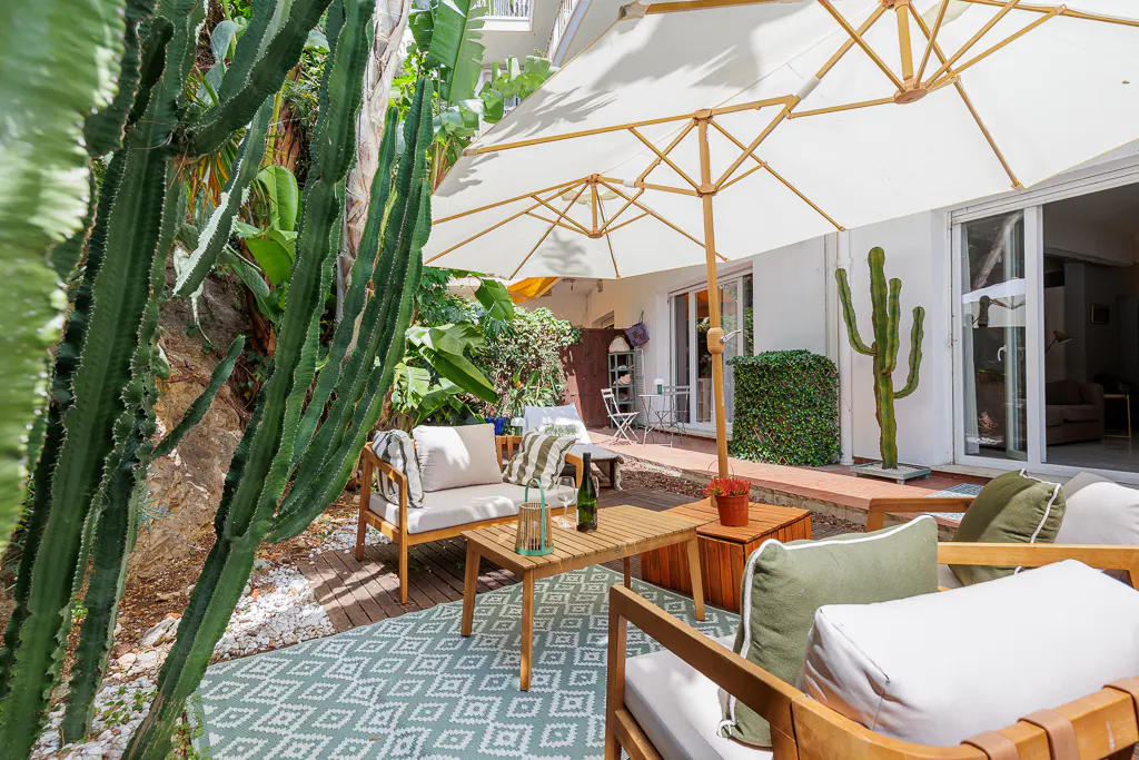 Outdoor patio with wooden furniture, white cushions, and a geometric rug. A large white umbrella provides shade, and cacti add a desert vibe.