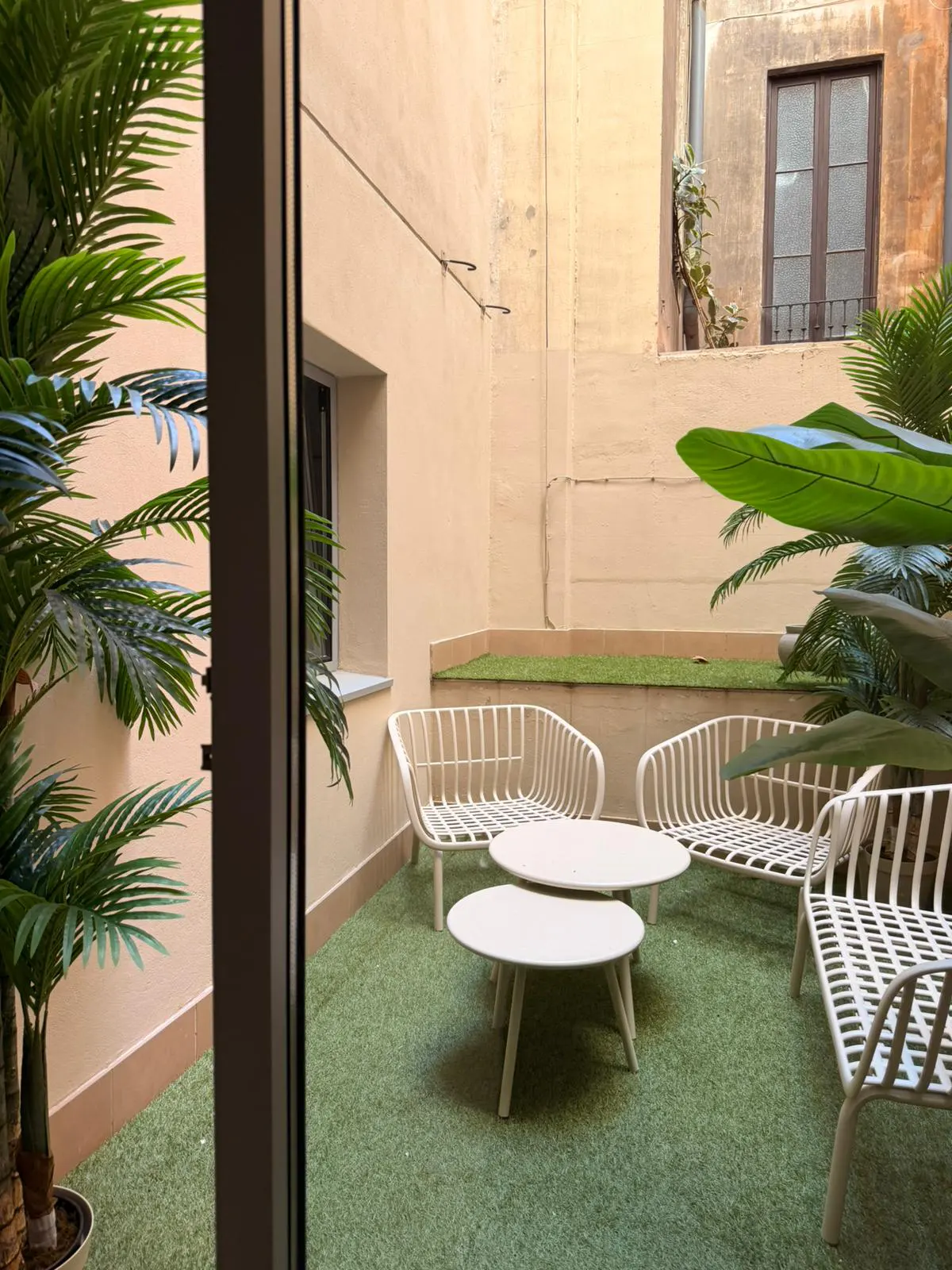 Outdoor patio with white metal furniture on green turf. Palm trees and building walls surround the space.