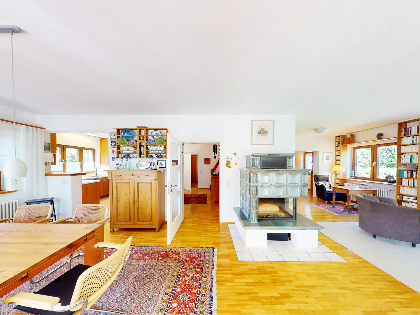 Bright, open-concept living space with wood floors. A dining table sits near a wooden cabinet and a tiled fireplace. Windows and bookshelves are visible.