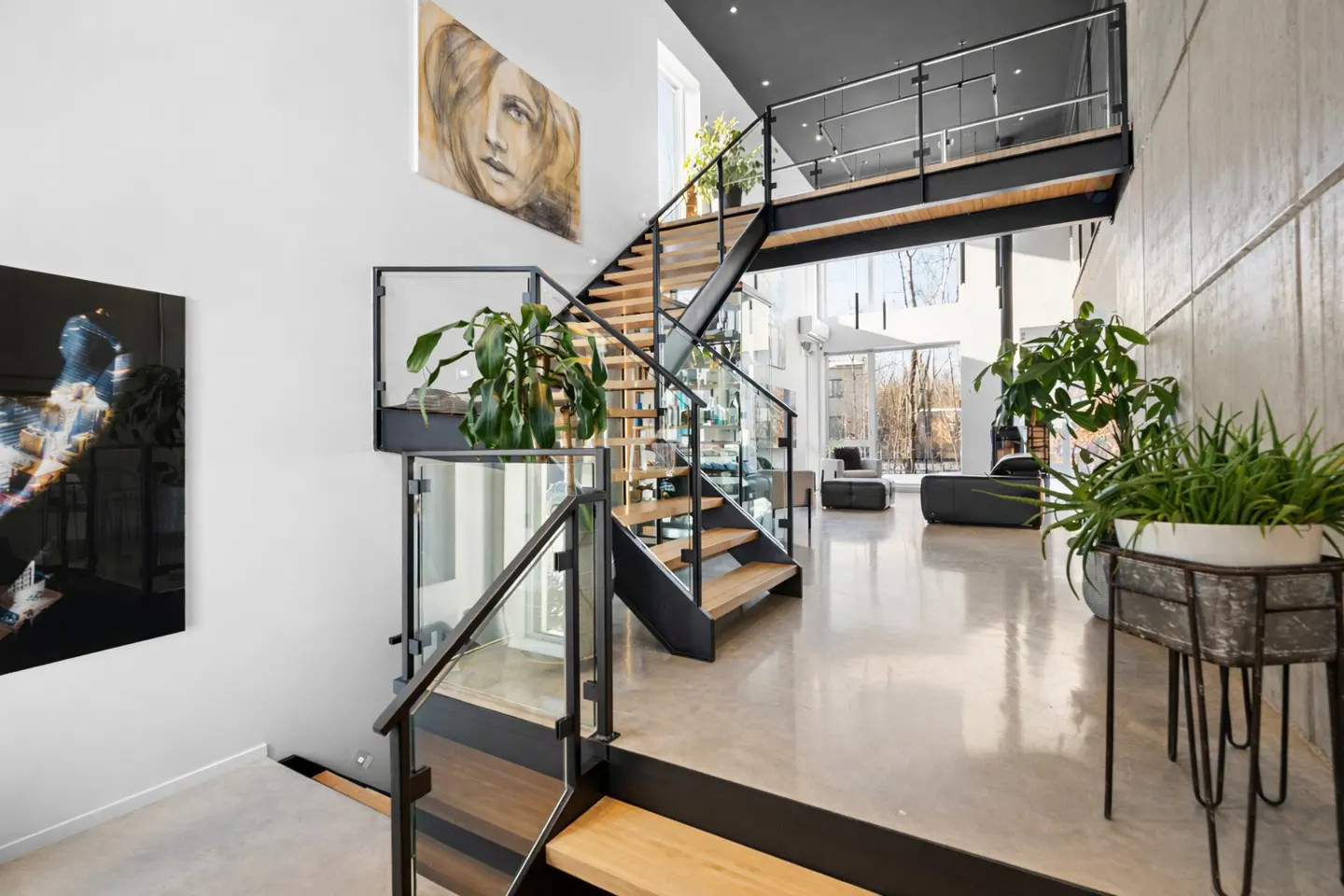Modern home interior with wooden stairs, glass railings, and black metal frames. Artwork adorns the white walls, and plants add greenery. Polished concrete floors reflect light.