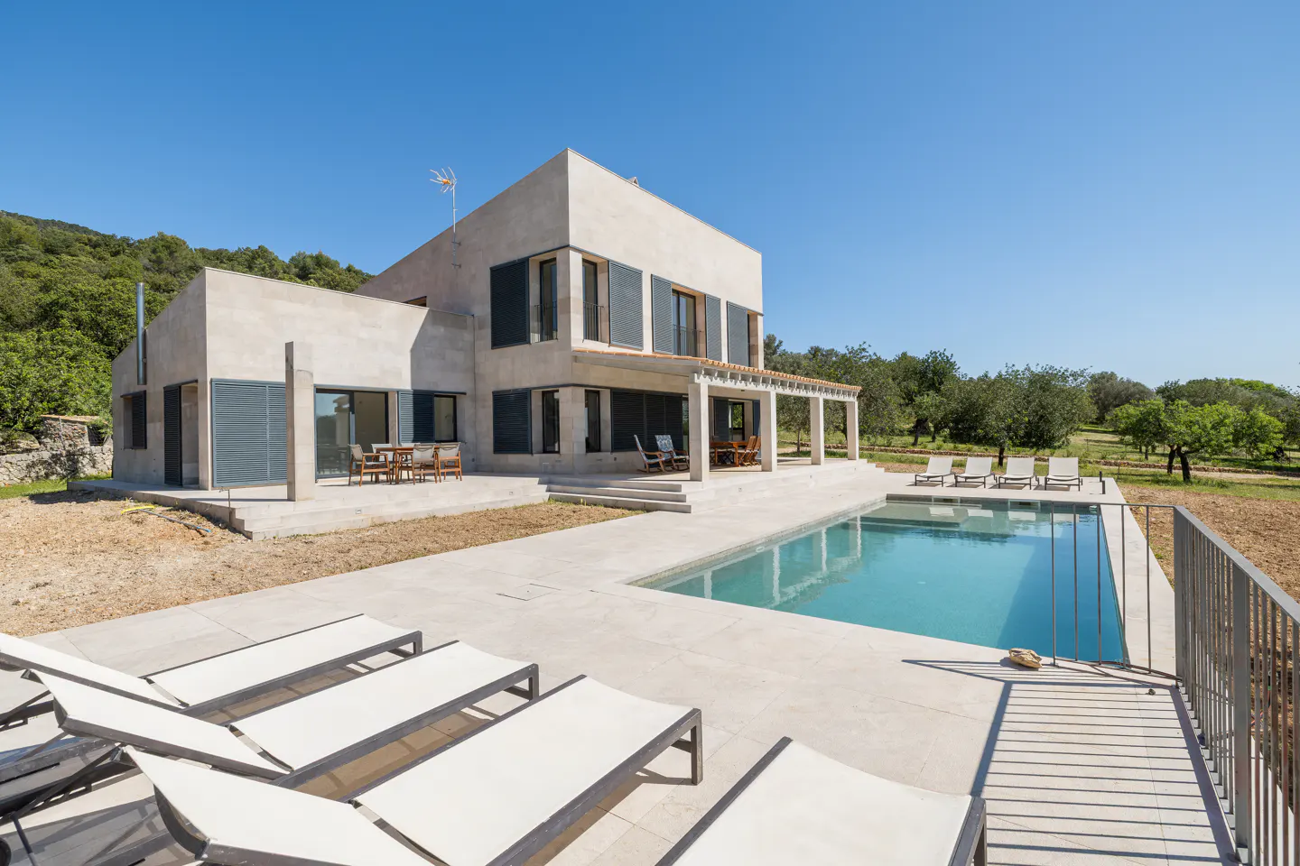 Modern two-story home with a pool. Lounge chairs sit in the foreground, with trees and a blue sky in the background.