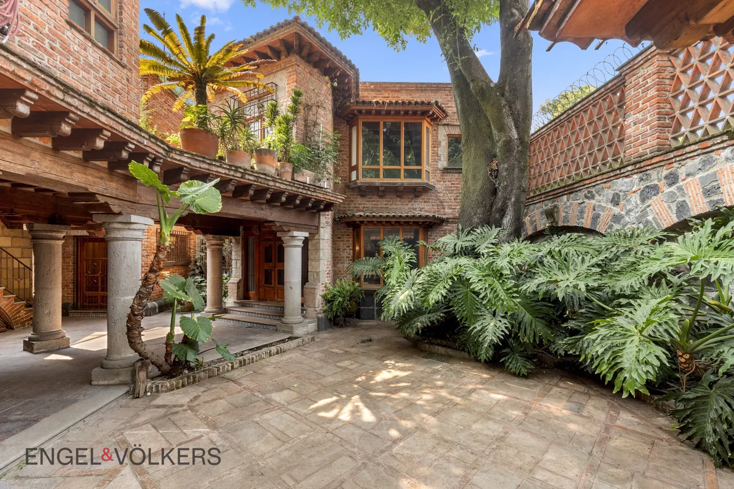 Courtyard view of a two-story brick house with a covered porch, columns, and lush greenery.
