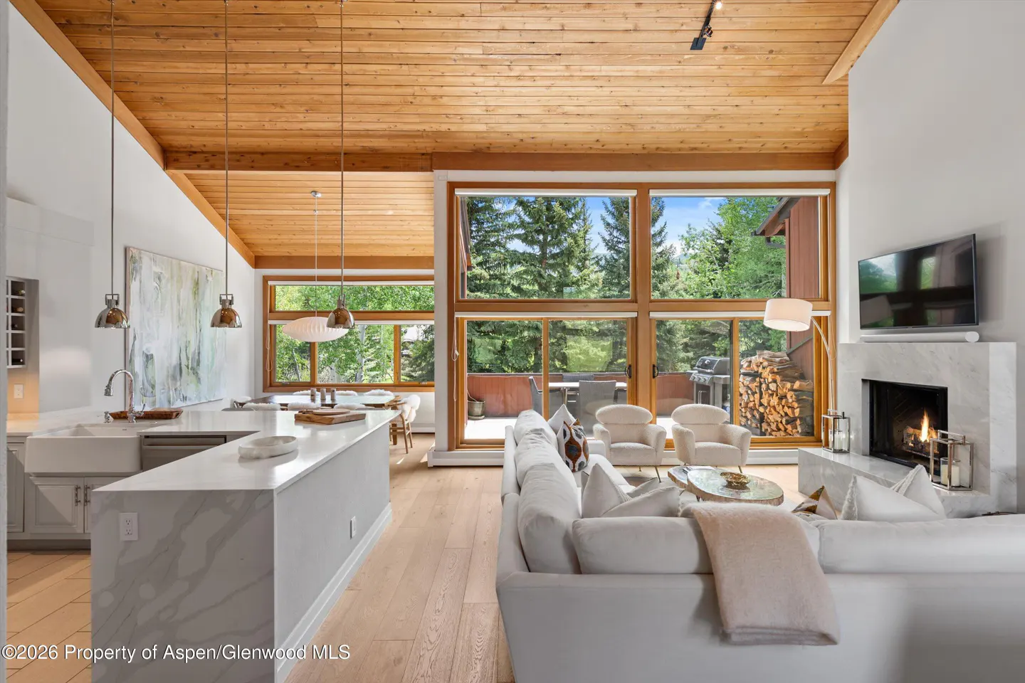 Modern open-concept living room with white sofa, marble fireplace, wood-paneled ceiling, and large windows facing trees.