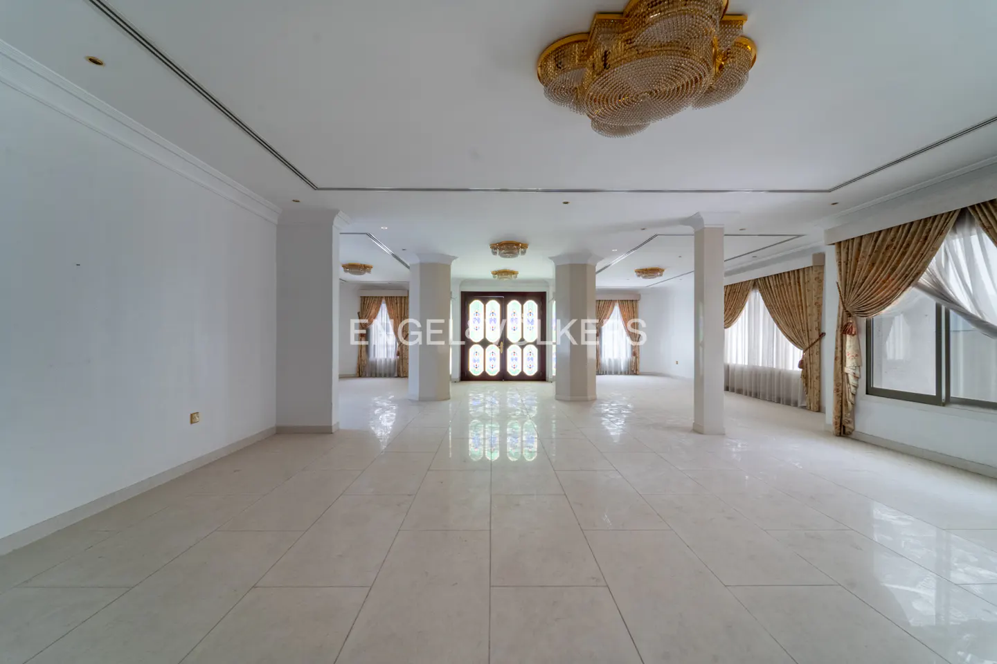 A spacious, empty room with white walls, marble floors, and gold chandeliers. Columns divide the space, and windows are draped with gold curtains.