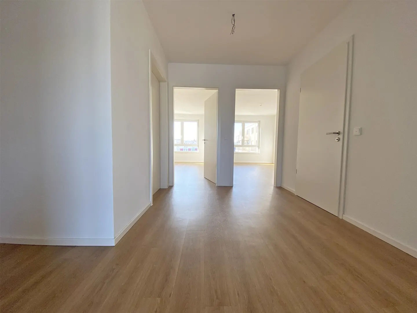 An interior shot of a bright, empty apartment hallway with light wood floors and white walls. Two open doorways lead to rooms with windows.