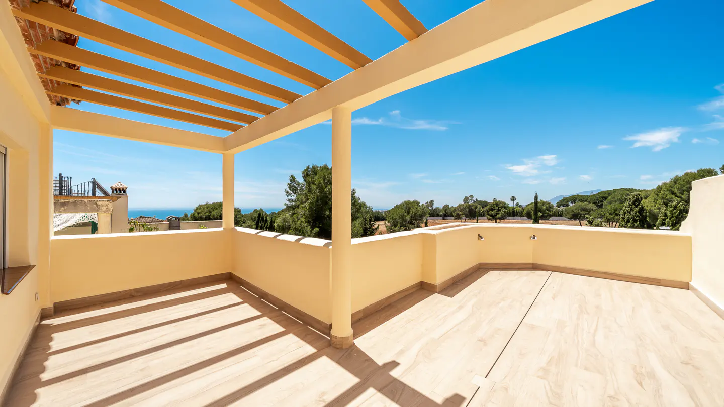 A sunny balcony with a beige pergola overlooks green trees and a blue sky. The floor is light beige tile, and shadows from the pergola create stripes.