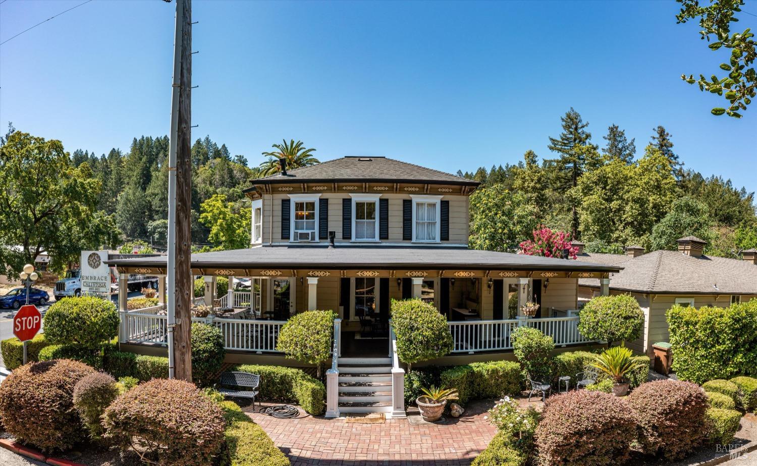 Two-story tan house with a wrap-around porch, white railings, and manicured landscaping under a clear blue sky.