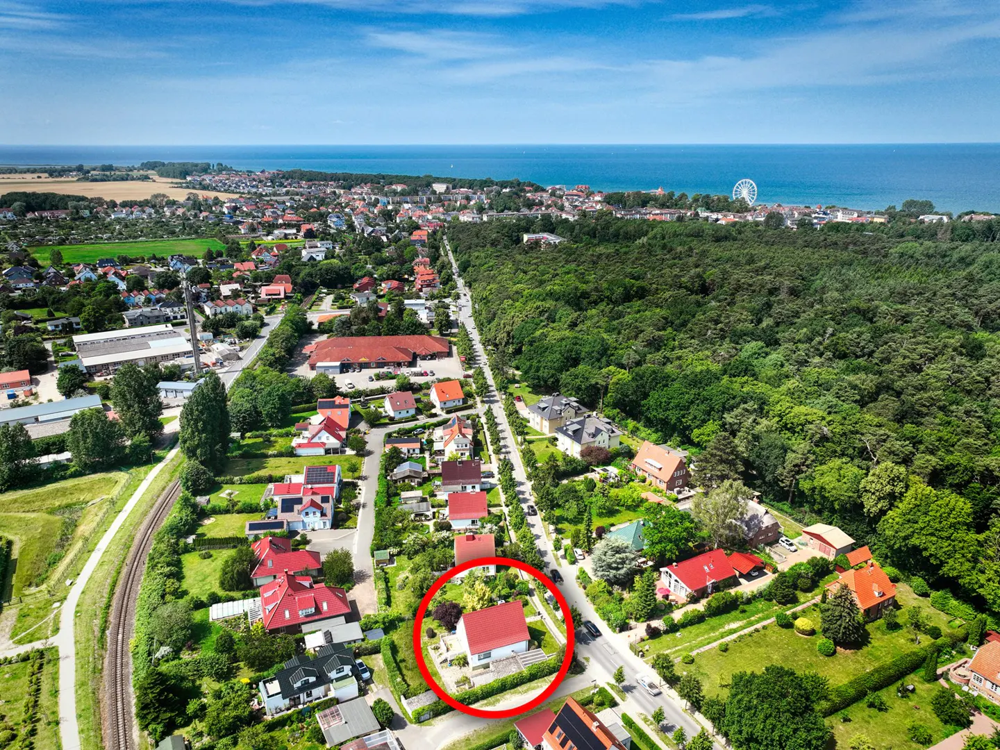 Aerial view of a red-roofed house circled in red, nestled in a green neighborhood near a forest and the ocean under a blue sky.