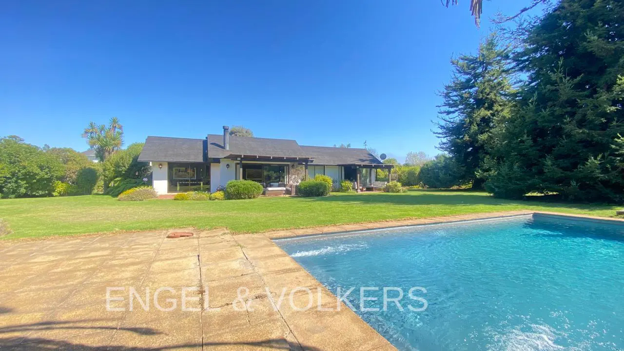 Exterior view of a white house with a black roof, green lawn, and a blue swimming pool on a sunny day.
