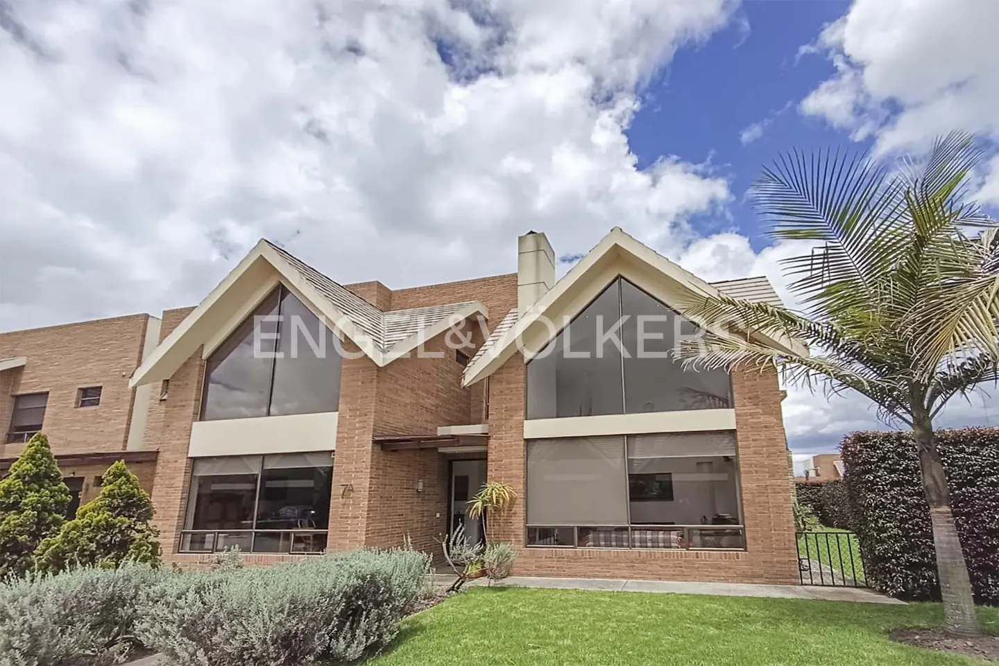 Two-story brick house with large windows and gabled roofs, set against a blue sky with clouds. Palm tree on the right. Engel & Volkers logo visible.
