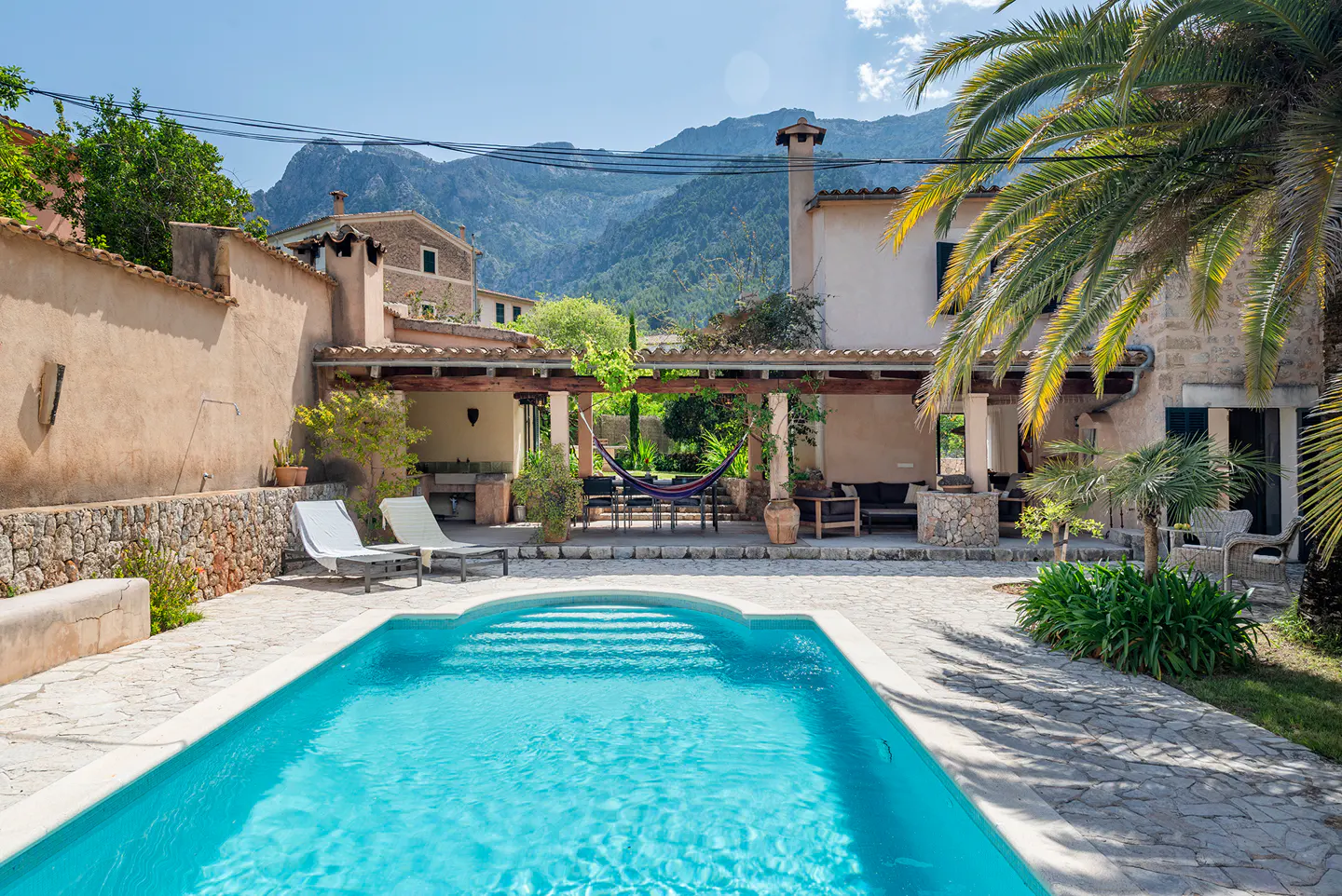 A turquoise pool with steps leads to a patio with a hammock, lounge chairs, and a stone house with a mountain backdrop.