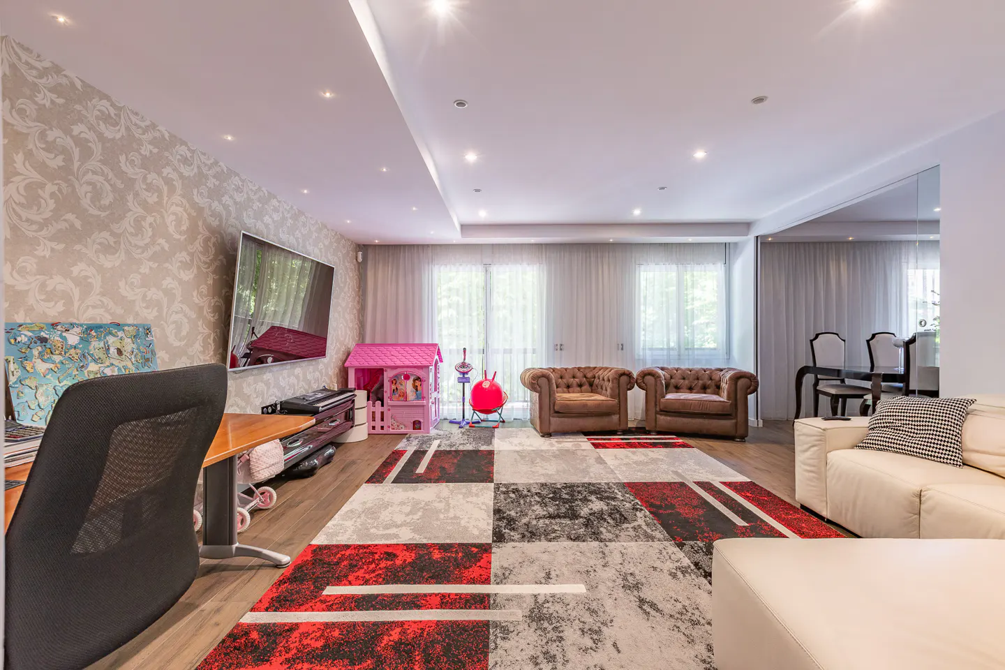 Bright living room with a desk, TV, pink playhouse, and brown leather chairs. A red, gray, and white rug covers the wood floor.