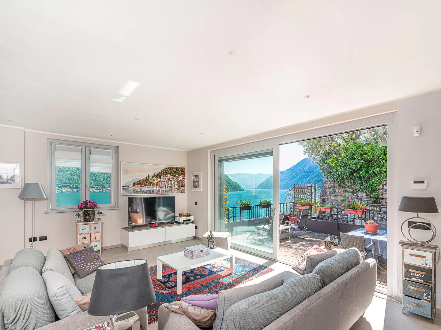 Bright living room with gray sofas, white table, and a view of a lake through sliding glass doors to a patio.