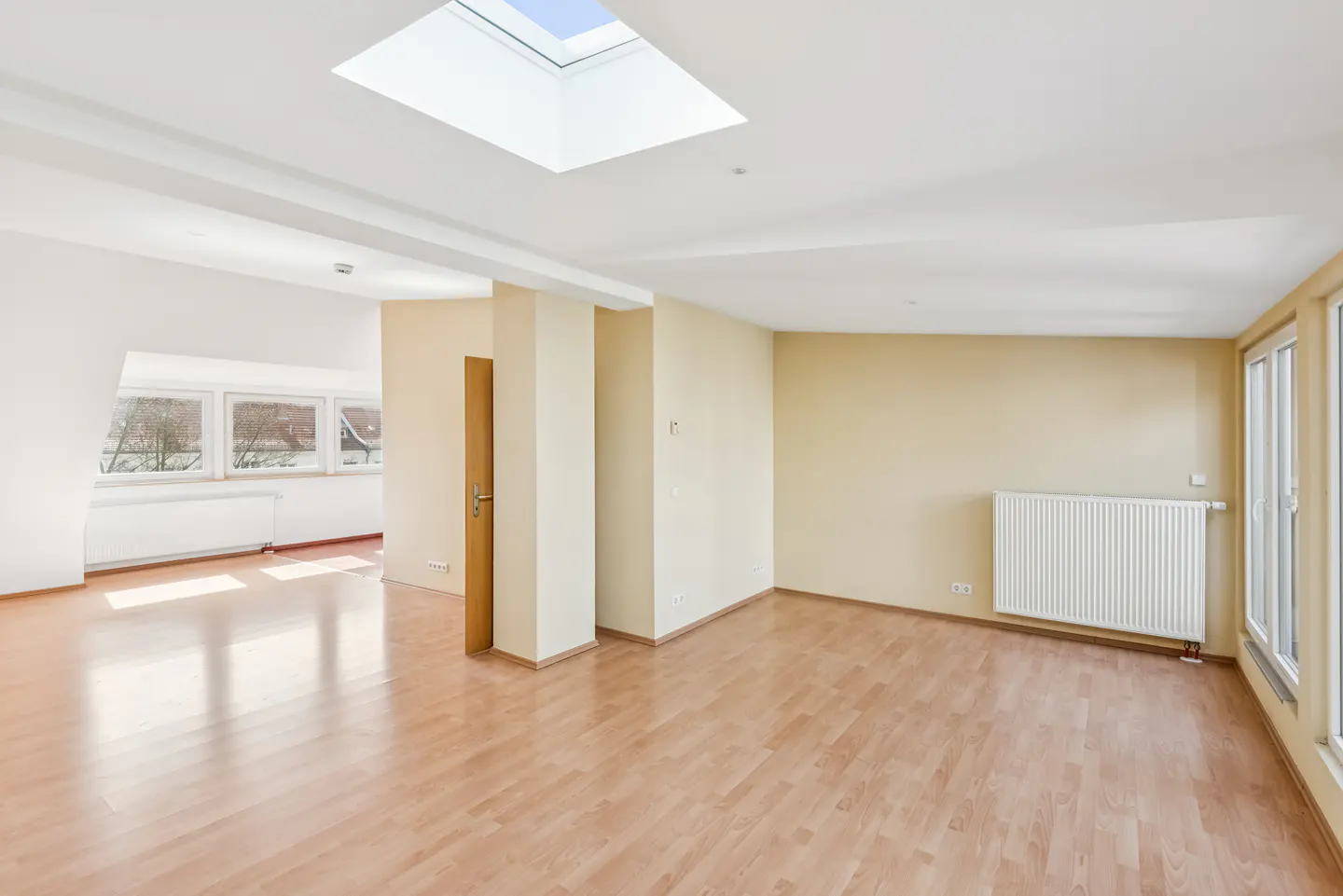 Bright, empty room with wood floors, beige walls, and a skylight. A doorway leads to another room with windows. A white radiator is visible.