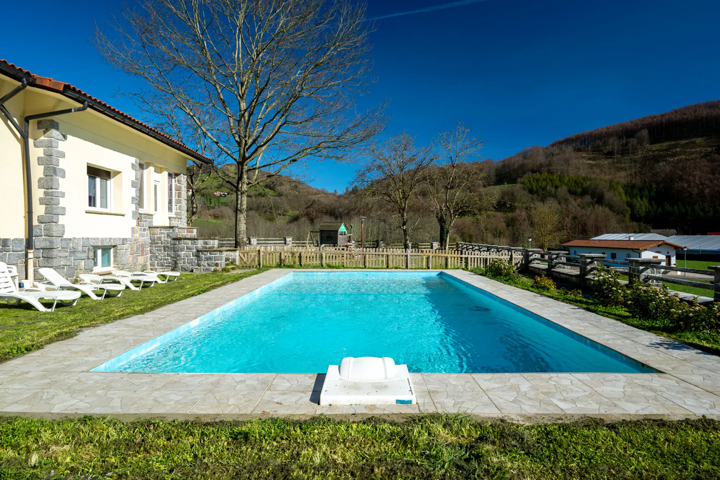 Rectangular swimming pool with blue water next to a cream-colored house, set against a backdrop of hills and blue sky.