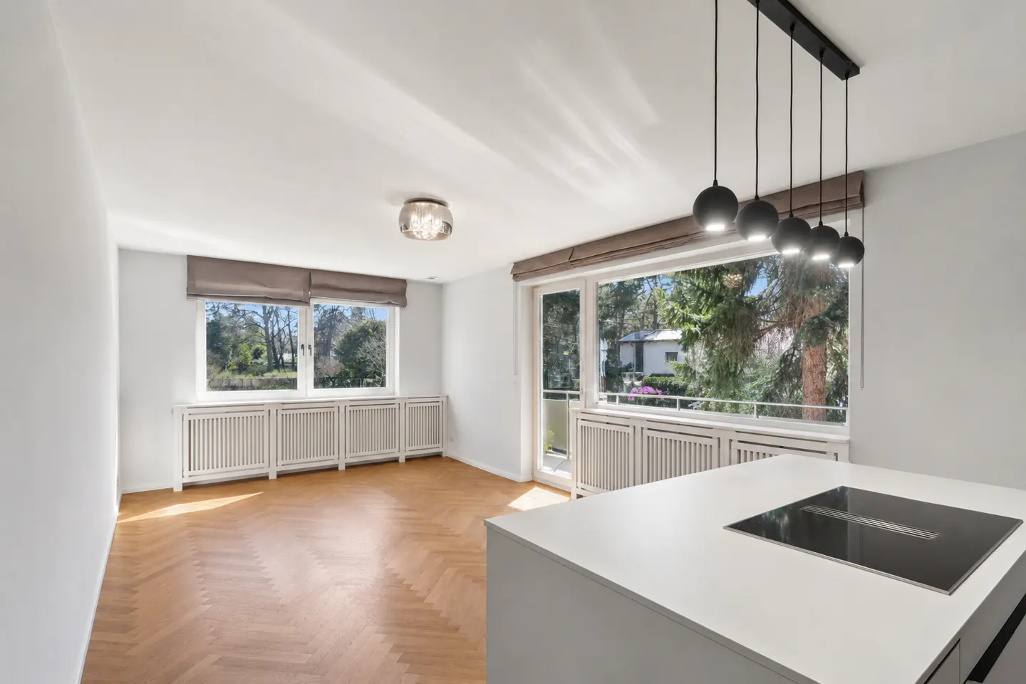 Bright, modern apartment interior with herringbone wood floors, white walls, and large windows overlooking trees. A white kitchen island with a cooktop is visible.