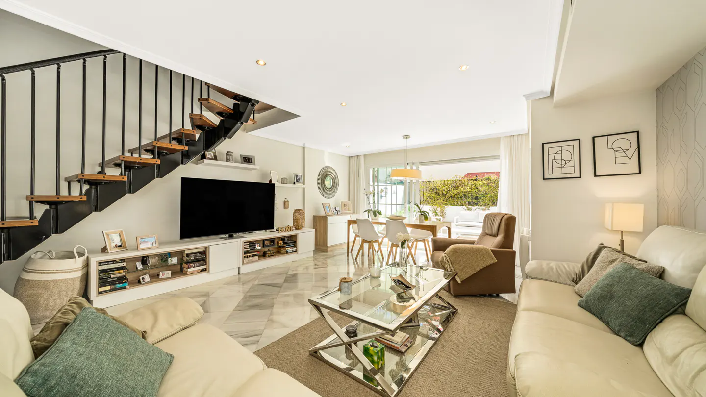 Bright living room with white sofas, glass coffee table, and dining area. Stairs with black railings lead up. Large TV on a white media console.