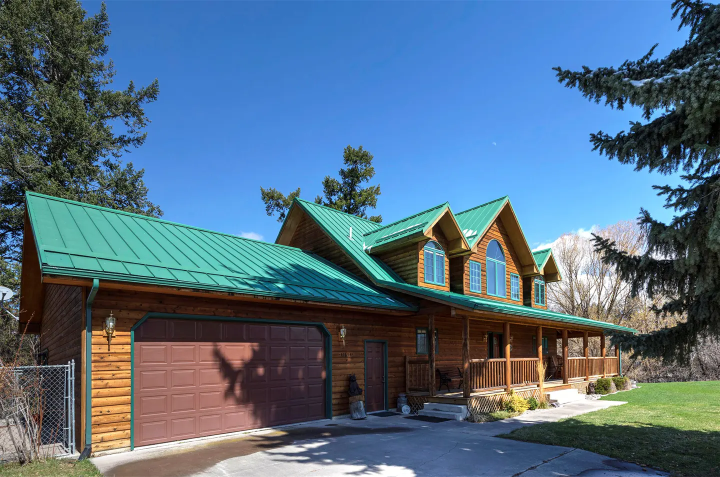 A log cabin home with a green metal roof, brown garage door, and a porch with wooden railings.