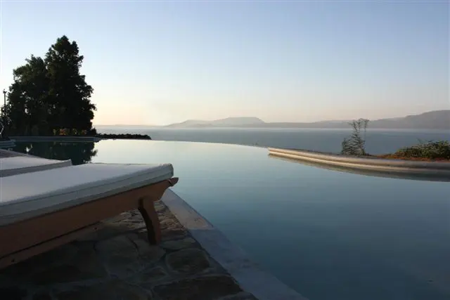 Infinity pool overlooking the ocean. A lounge chair sits on a stone patio. Trees and mountains are visible in the distance.