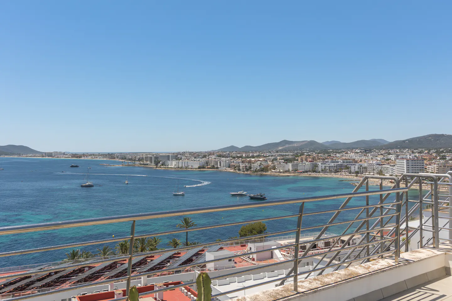 View from a balcony overlooking a blue bay with boats, a sandy beach, and a coastal town under a clear blue sky.