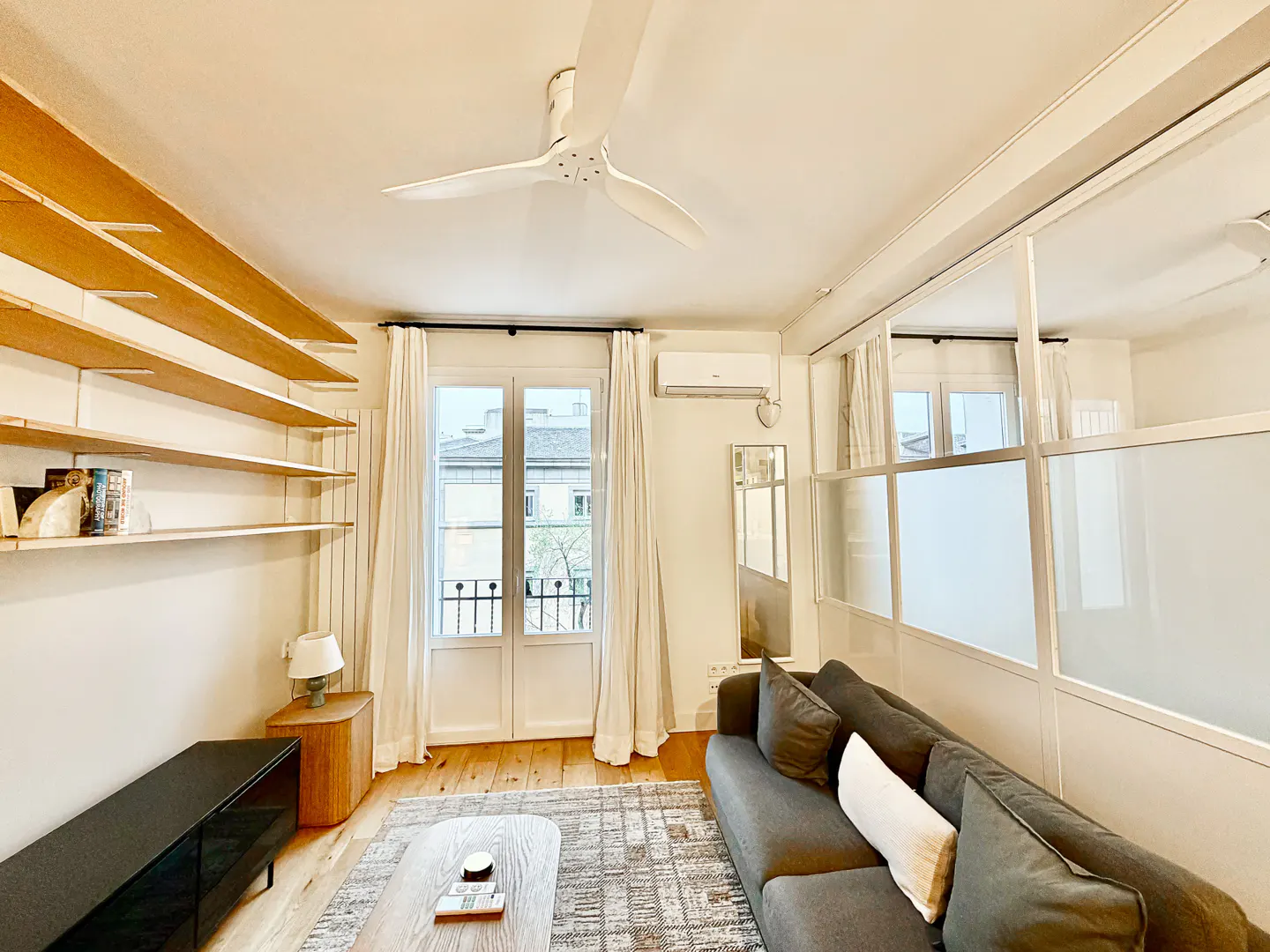 Bright living room with a gray sofa, wood floors, and white walls. Shelves line one wall, and a window with white curtains overlooks a city street.