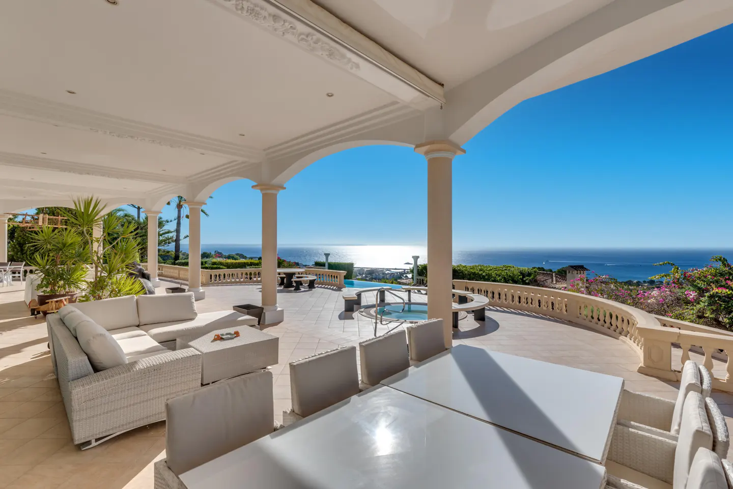 Luxury patio with white furniture, columns, and arches overlooking the blue ocean on a sunny day.