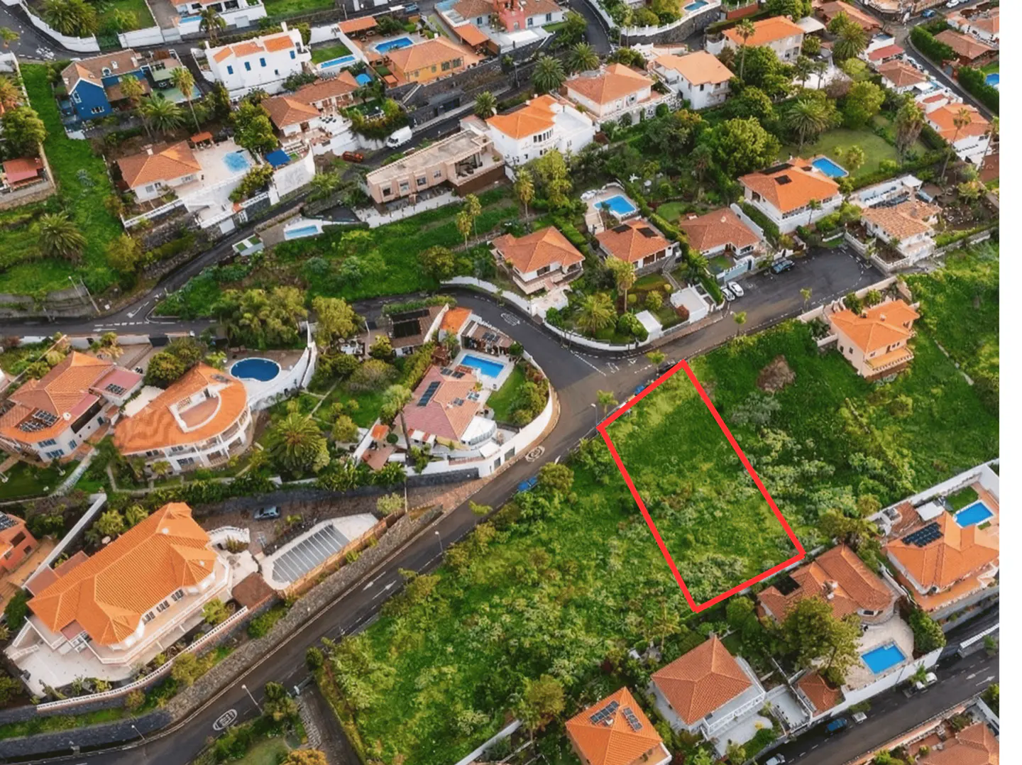 Aerial view of a grassy lot outlined in red, surrounded by houses with orange roofs and green trees.