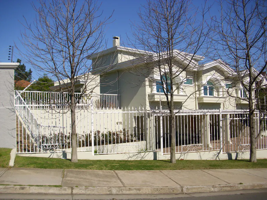 Two-story beige house with white trim and a white metal fence, viewed from the street on a sunny day.