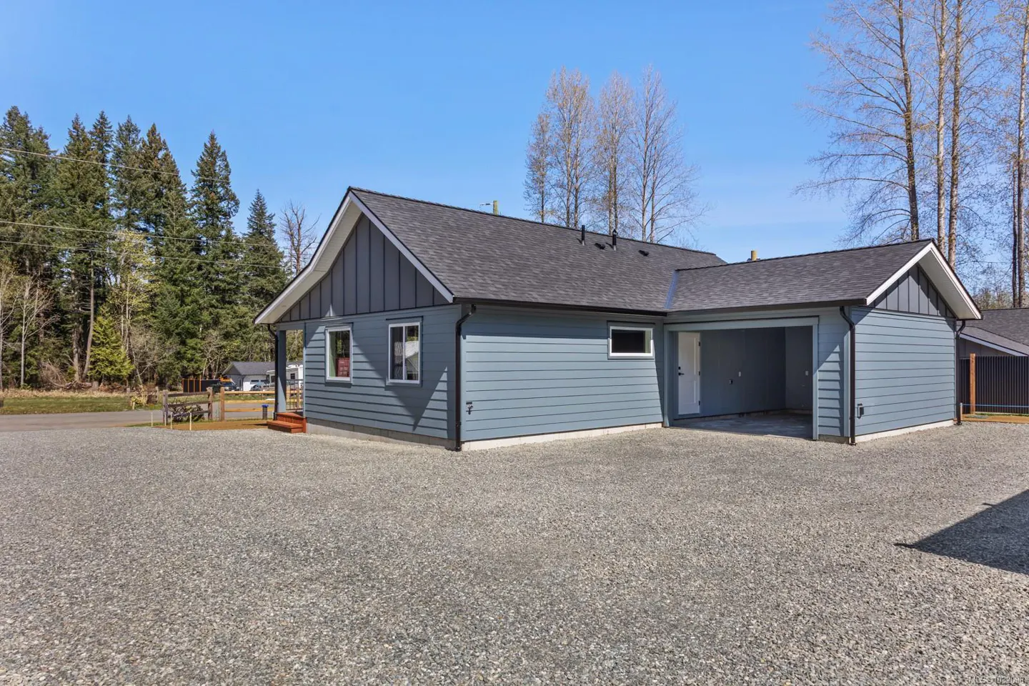 Exterior view of a blue, single-story house with a dark gray roof and a gravel driveway on a sunny day.