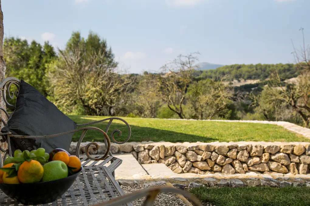 Outdoor patio view with a metal bench, black pillow, and fruit bowl. A stone wall borders a green lawn with trees and hills in the background.