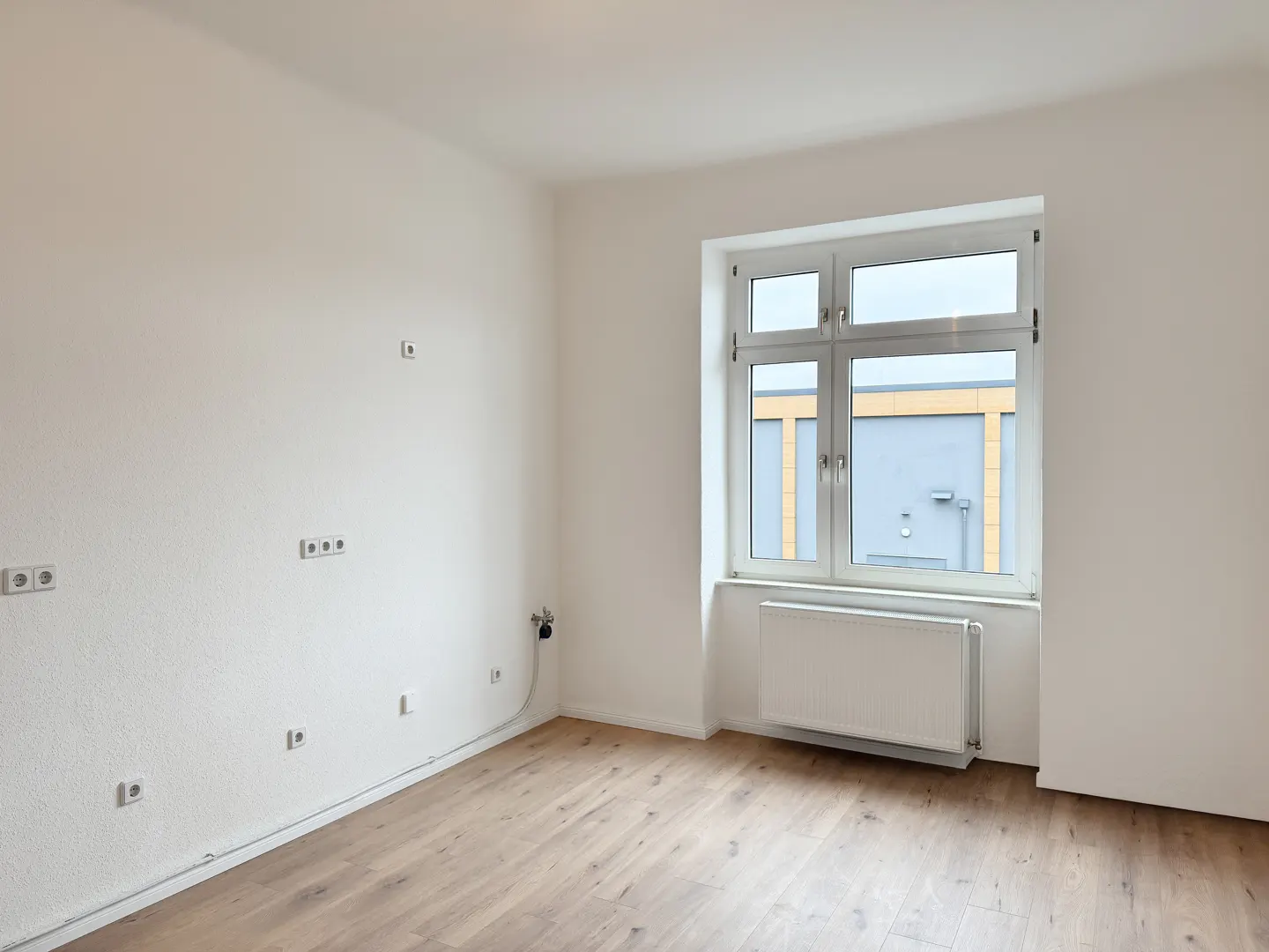 Bright, empty room with light wood floors, white walls, and a window. Electrical outlets are visible on the left wall. A white radiator sits under the window.