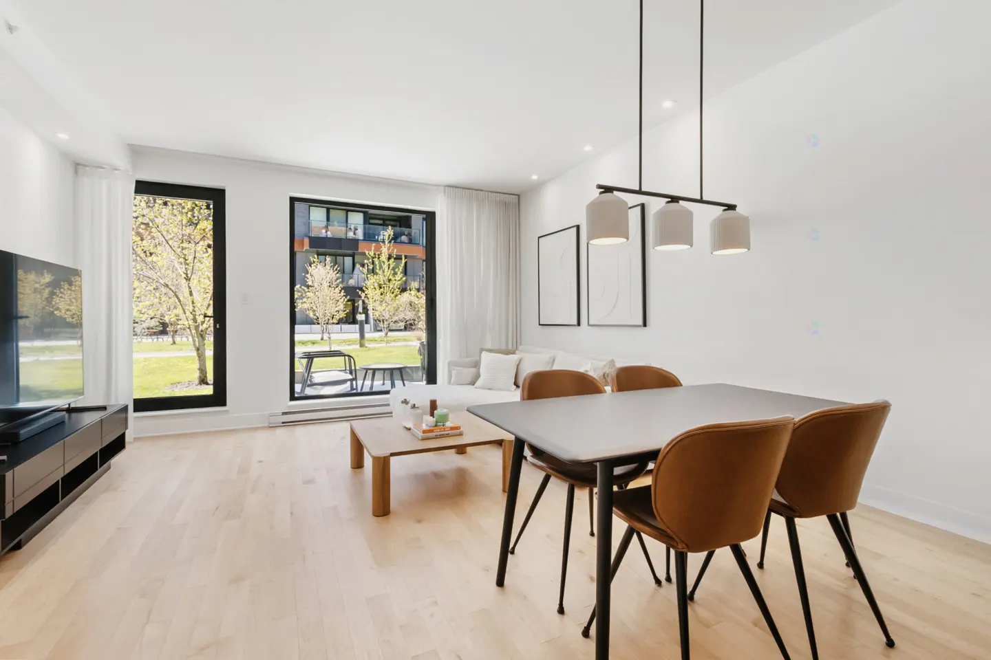 Bright, modern living room with wood floors, a dining table with brown chairs, and a view of a green lawn through large windows.