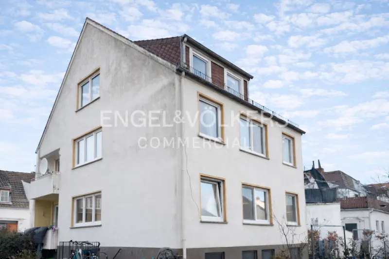 Exterior of a three-story white building with brown trim and a red tile roof under a cloudy blue sky. "Engel & Völkers Commercial" is printed on the building.