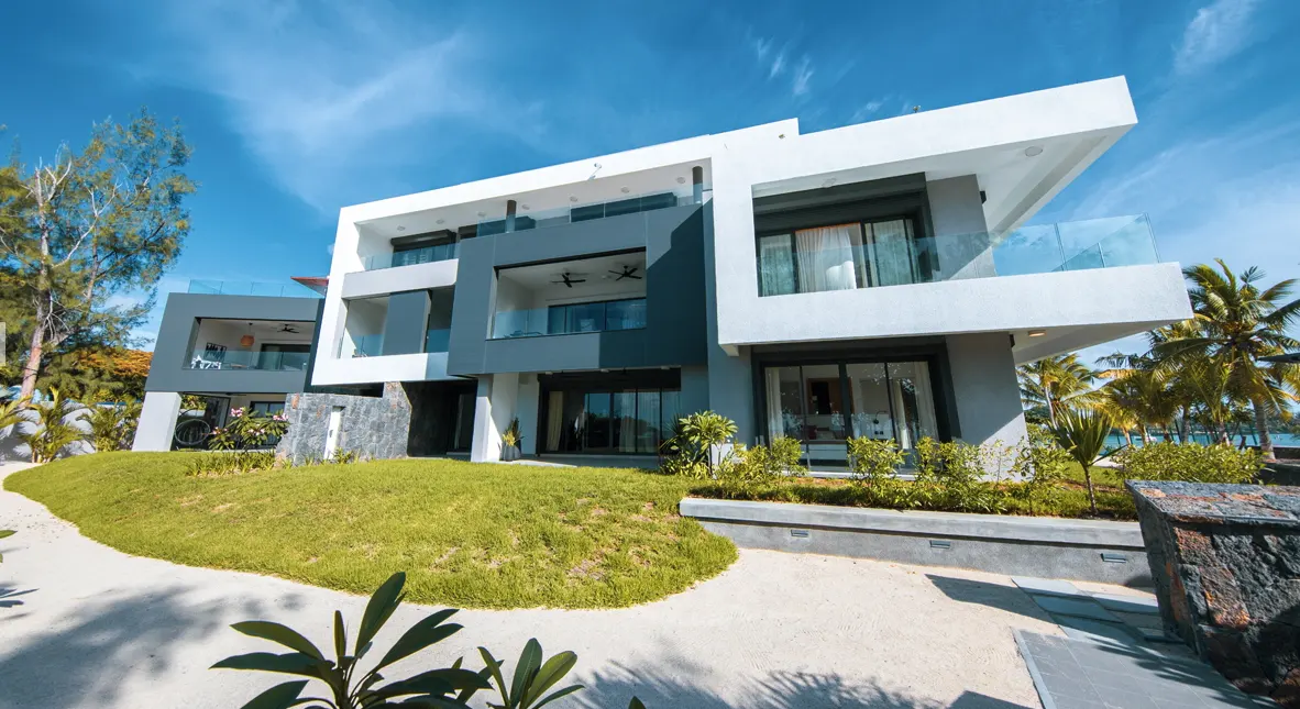 Modern, multi-story home with white and gray exterior, glass balconies, and large windows. Lush green lawn and tropical trees surround the property.