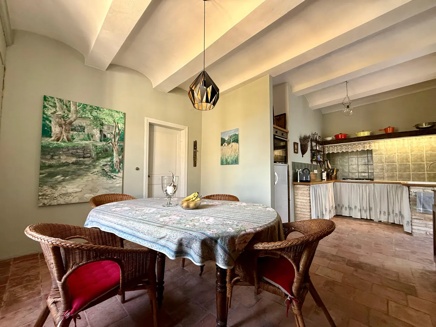 Dining area with a table, wicker chairs with red cushions, and a kitchen in the background. The walls are light green.