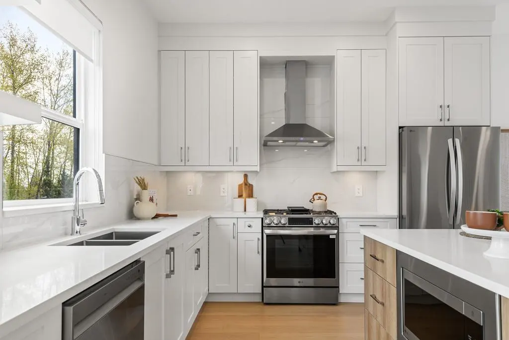 Bright, modern kitchen with white cabinets, stainless steel appliances, and a large window overlooking trees.