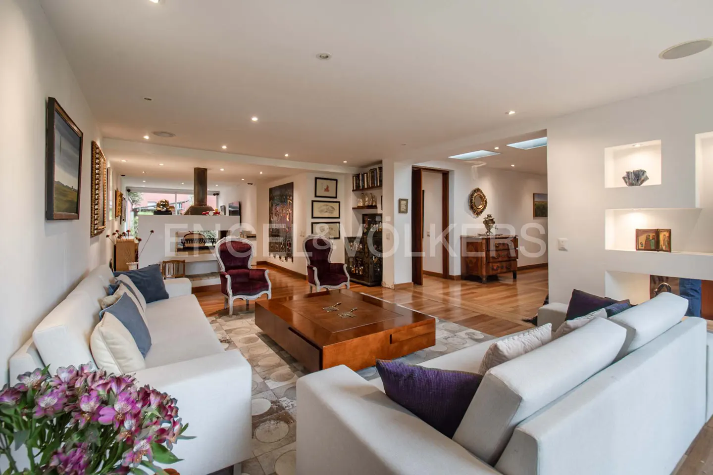 Open-concept living room with white sofas, purple accents, and a large wooden coffee table on a patterned rug. Hardwood floors and recessed lighting.