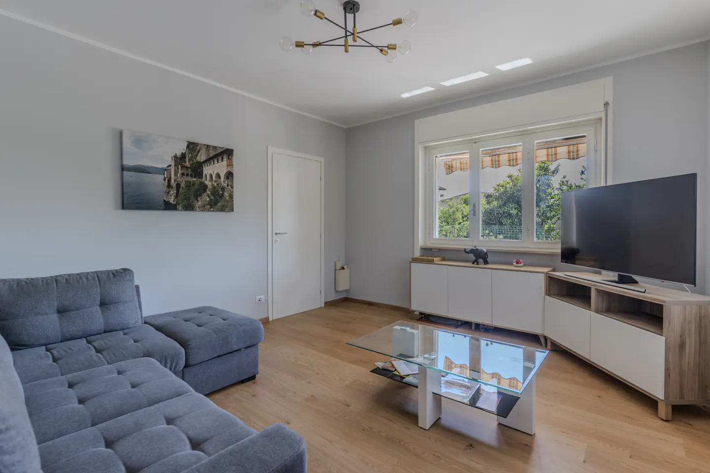 Living room with gray walls, wood floors, and a gray sectional sofa. A glass coffee table sits in front of a TV and white cabinets. A picture hangs on the wall.