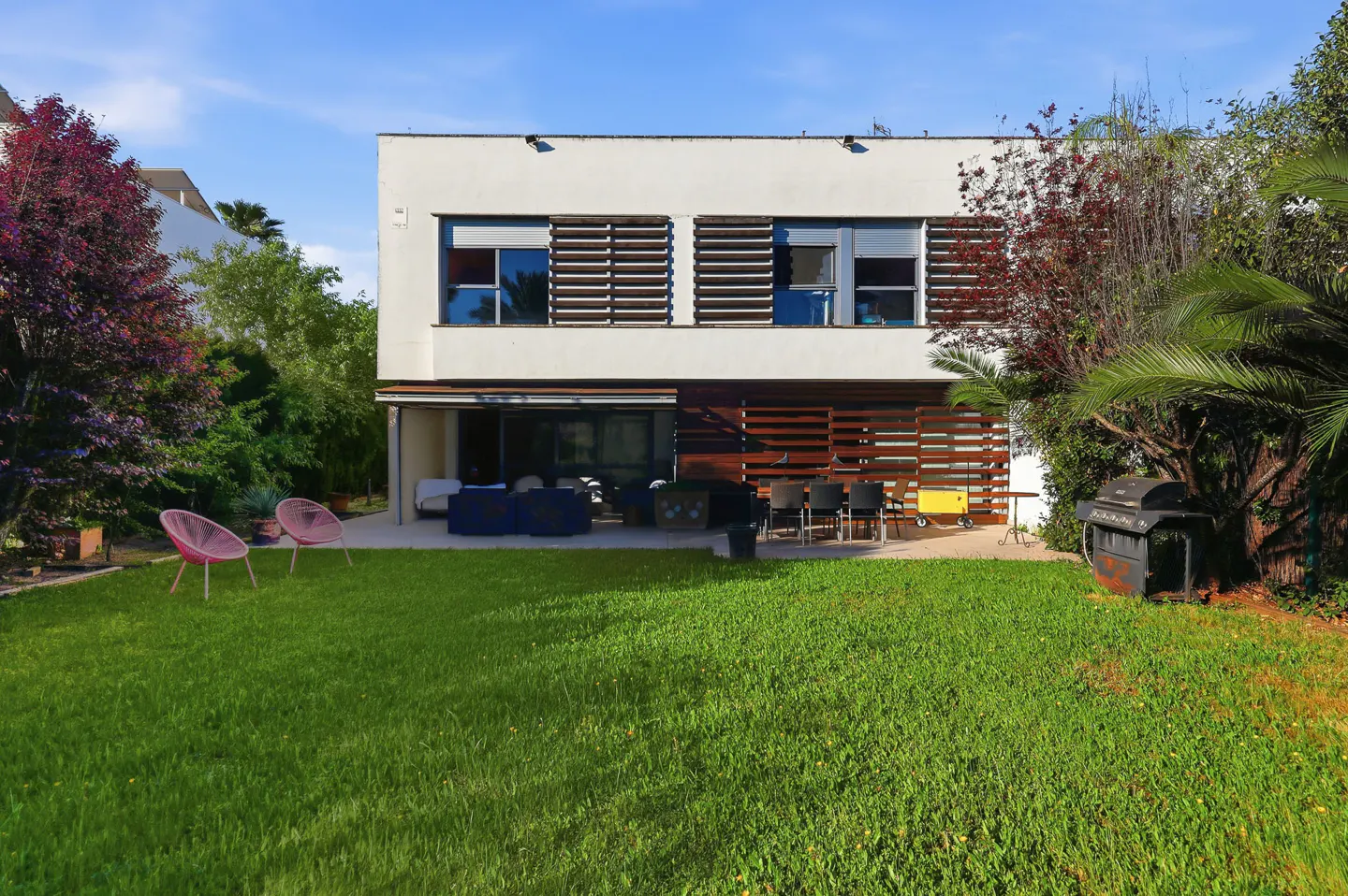 Two-story white house with a green lawn, patio furniture, grill, and trees under a blue sky.