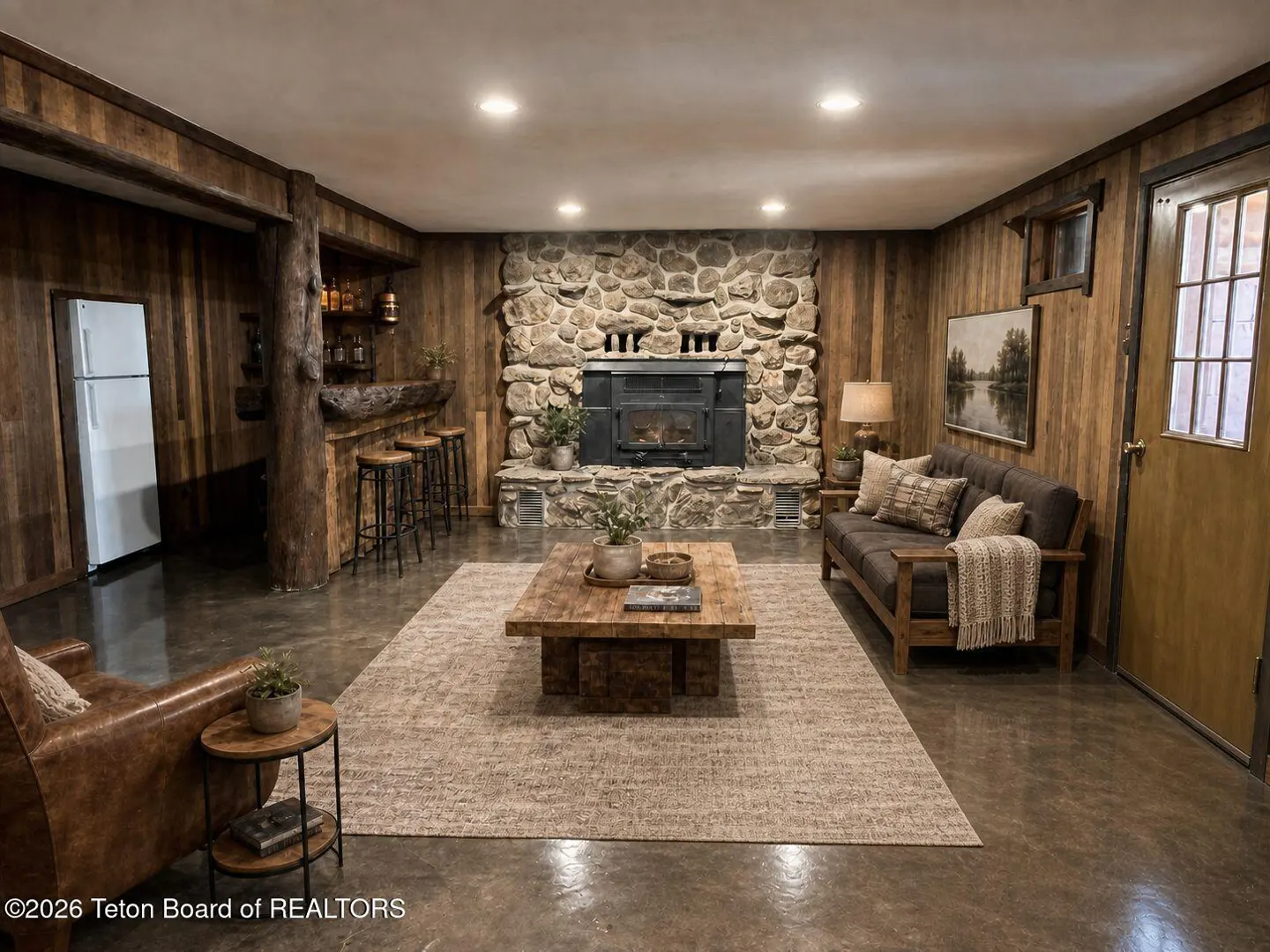 Rustic living room with wood paneled walls, stone fireplace, wooden bar, and furniture on a large area rug.