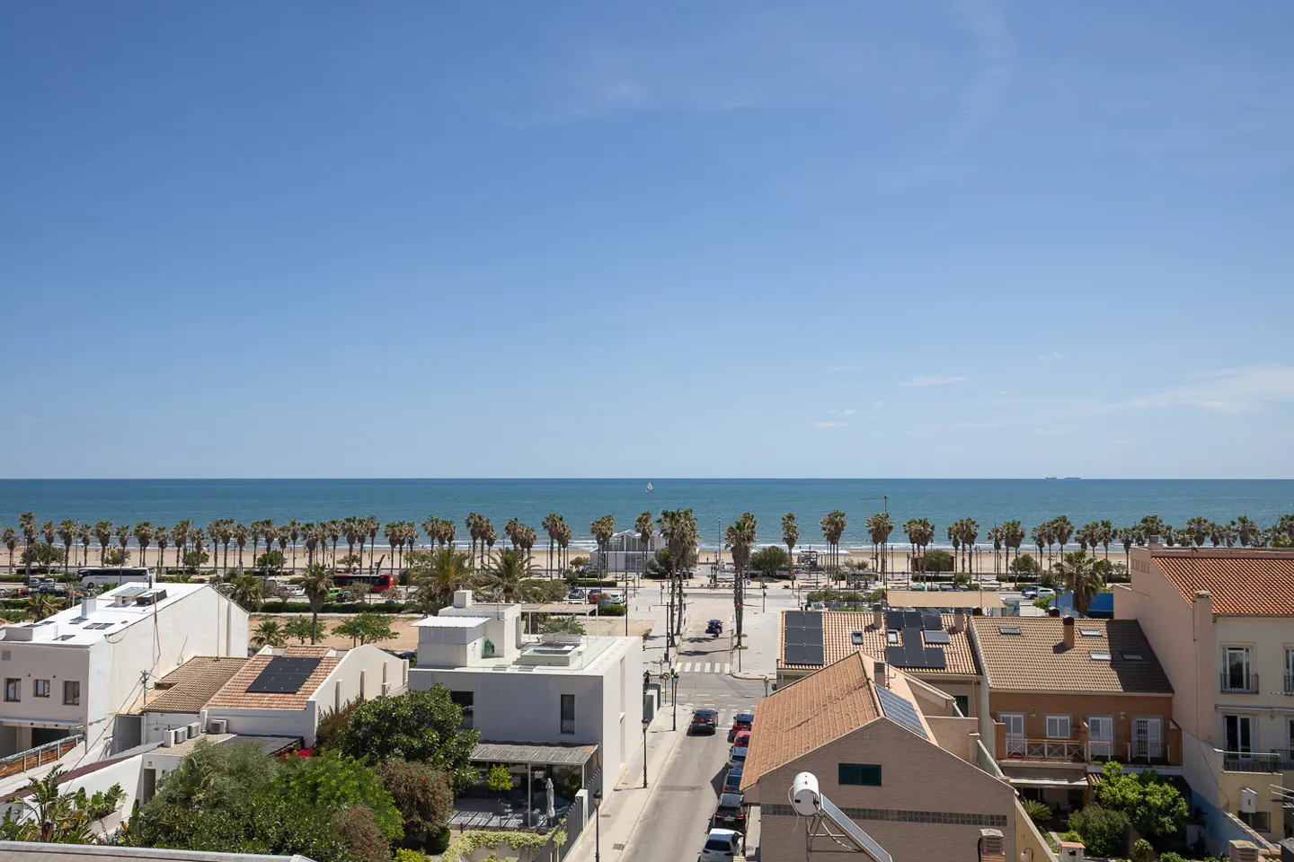 View of a coastal town with palm trees lining the beach, blue sky, and houses with solar panels.