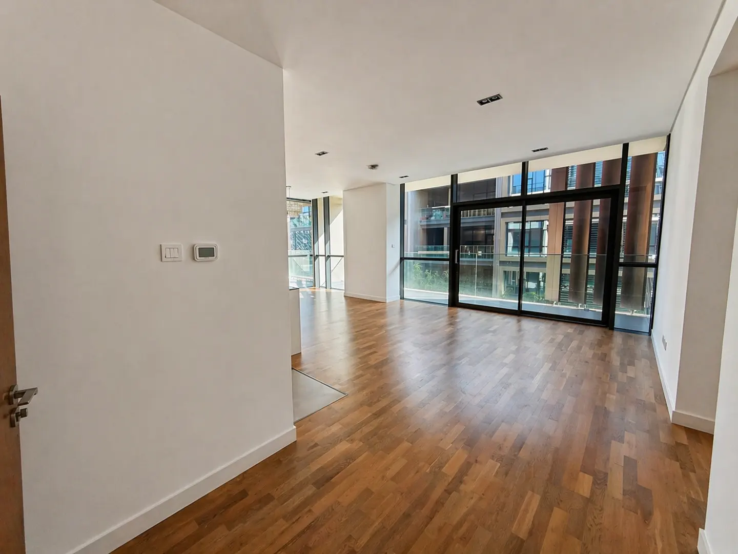 Bright, empty room with wood floors and white walls. Large windows show a building exterior. A thermostat is on the wall.