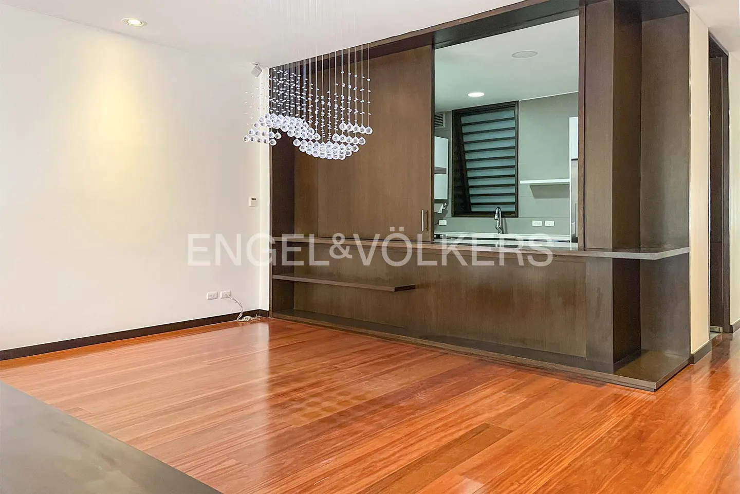 A modern living room with hardwood floors, a crystal chandelier, and a view into the kitchen through a dark wood partition.