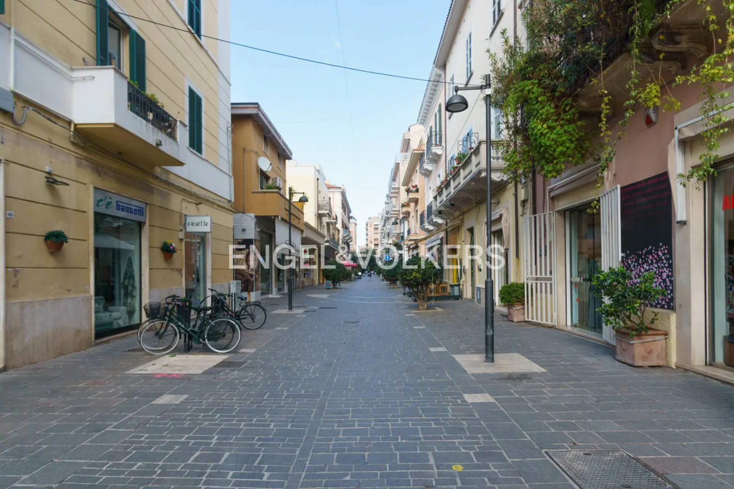 A narrow, paved pedestrian street in an Italian town lined with multi-story buildings, storefronts, and potted plants.