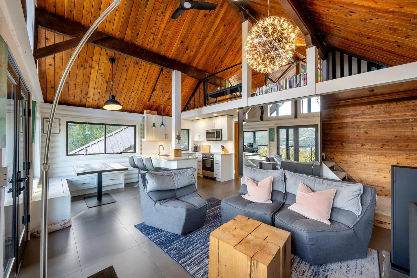 Open-concept living space with wood-paneled ceiling, gray seating, and a modern kitchen. A unique light fixture hangs above.