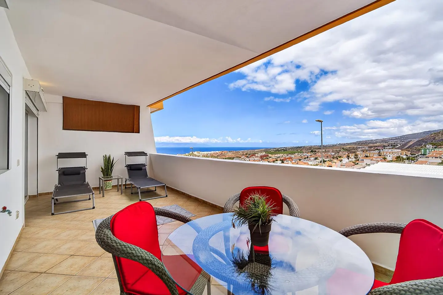 Balcony with a glass table, red chairs, and lounge chairs overlooking a coastal town and ocean under a blue, cloudy sky.