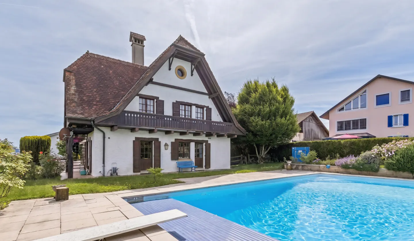 Exterior view of a white house with brown shutters and a red tile roof, next to a blue swimming pool on a sunny day.